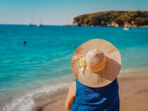 Вид на молодую женщину в соломенной шляпе и синем платье, отдыхающую на пляже и смотрящую вперед в лагуну в Парга Греция
. - Фото, изображение