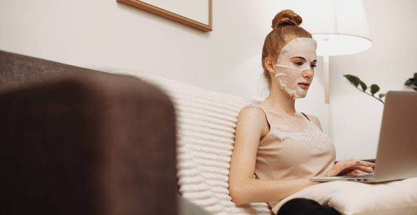 Red haired caucasian woman applying a paper mask on her face while sitting on the sofa with a computer - 写真・画像