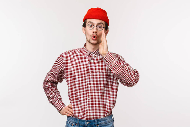 Waist-up portrait of funny young guy with beard and moustache in red beanie, glasses, calling someone, searching for friend in crowd, hold hands near mouth gossiping, whisper secret - Photo, Image