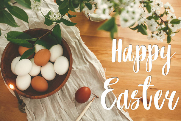 Happy Easter text. Easter greetings lettering. Natural eggs in wooden bowl and cherry blooming flowers on rustic wooden table. Greeting card. - 写真・画像