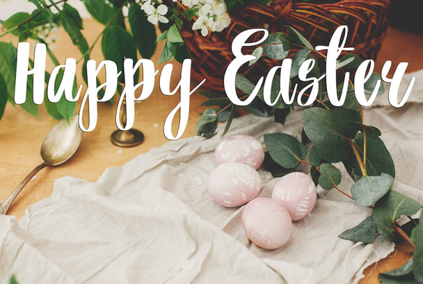 Happy Easter text. Easter greetings lettering. Modern Easter eggs with eucalyptus branch on rustic wooden table with basket and candle. Greeting card - Photo, Image