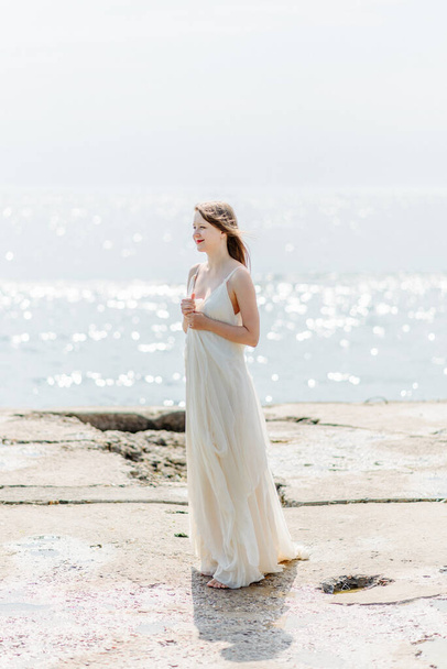 Een jong mooi meisje in een lange melkkleurige jurk loopt langs het strand en de pier tegen de achtergrond van de zee. - Foto, afbeelding