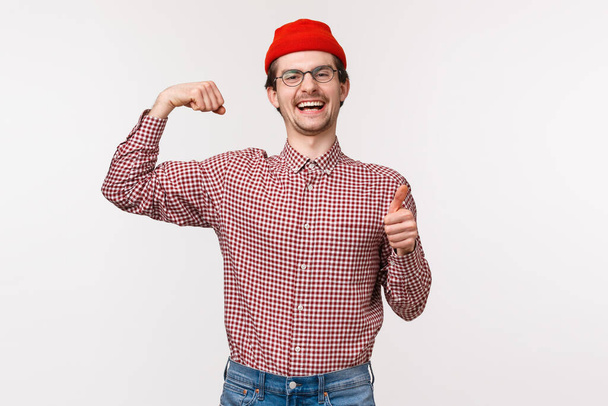 Vita-up ritratto infastidito uomo felice e allegro in berretto rosso, occhiali, mostrare pollici in su e bicipiti flessibili come vantarsi di quanto sia forte, fiducioso con il suo corpo, in piedi sfondo bianco
 - Foto, immagini