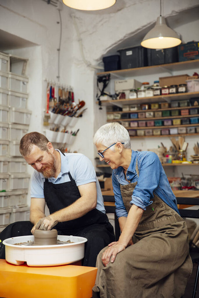 Mujer mayor girando arcilla en una rueda con la ayuda de un profesor en la clase de cerámica - Foto, imagen