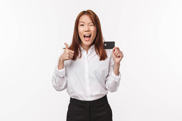 Retrato de satisfeito e confiante mulher asiática de boa aparência na camisa piscar a câmera satisfeito, mostrar polegares para cima e cartão de crédito, recomendo serviço bancário, encantado com qualidade, fundo branco
 - Foto, Imagem