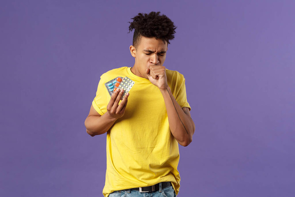 Health, influenza, covid-19 concept. Portrait of ill young man caught cold, have severe caugh, holding pills, showing drugs bought in drugstore, standing purple background feeling bad - Photo, Image