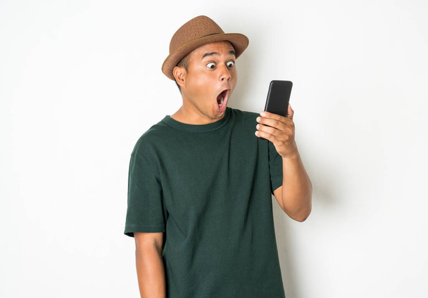 Азиатские мужчины используют смартфоны. Он смотрит на телефон, потом делает очень удивленное лицо, надевает старую зеленую рубашку и носит коричневую шляпу. Ему около 27-35 лет.
. - Фото, изображение