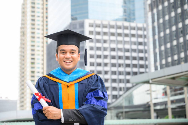 Gelukkige afstuderen. Happy Asain man in diploma toga 's met diploma in de hand op stedelijke achtergrond. - Foto, afbeelding