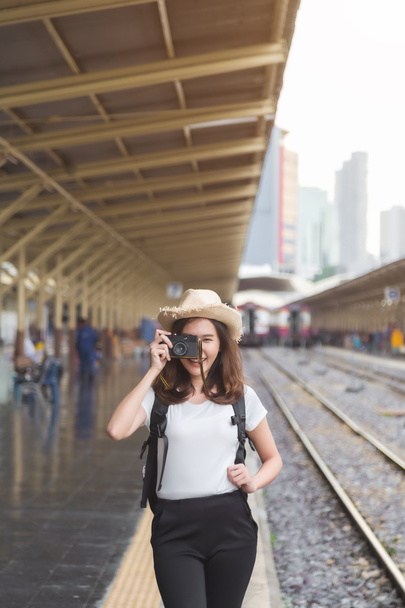 Średnie zbliżenie piękna młoda Azjatka podróżnik w białym t-shirt i plecak celuje aparat fotograficzny i uśmiech stojąc obok kolei na stacji kolejowej. - Zdjęcie, obraz