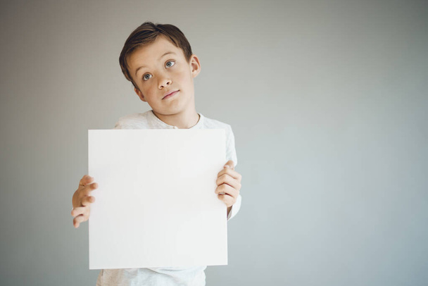cool young boy holding white empty sheet in front of grey background  - Φωτογραφία, εικόνα