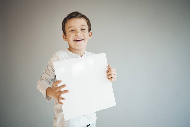 cool young boy holding white empty sheet in front of grey background  - Valokuva, kuva