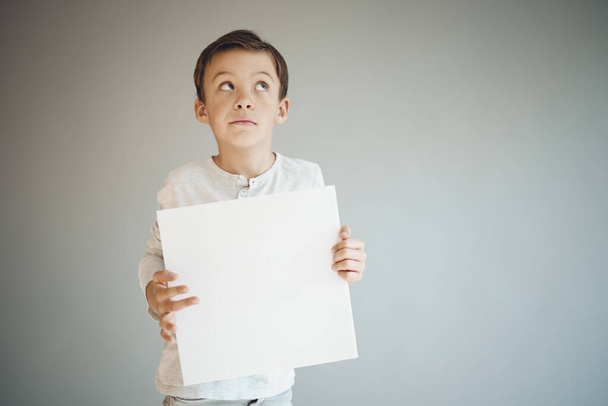 cool young boy holding white empty sheet in front of grey background  - Foto, afbeelding