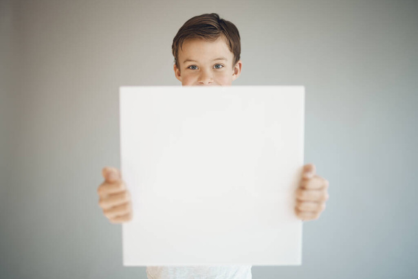 cool young boy holding white empty sheet in front of grey background  - Photo, Image