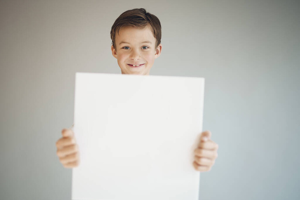 cool young boy holding white empty sheet in front of grey background  - Foto, Imagen