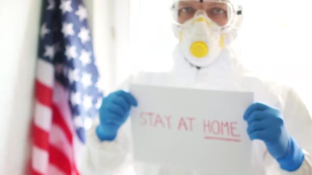 American doctor in a protective suit and mask on the background of the US state flag. A man is holding a picture with the inscription stay at home. Coronovirus covid19 and quarantine in America - Imágenes, Vídeo