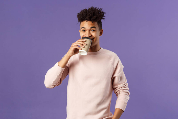 Freizeit-, Lifestyle- und Personenkonzept. Porträt eines jungen, fröhlichen Mannes, der Kaffee zum Mitnehmen aus seinem Lieblingscafé trinkt, lächelnd einen schönen Frühlingstag genießt und lila Hintergrund hat - Foto, Bild