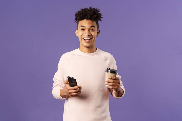 Waist-up portrait of happy enthusiastic hispanic man with smartphone, hold mobile phone and drinking coffee from take-away cup in office cafe, discuss recent project coworkers and team - Photo, image