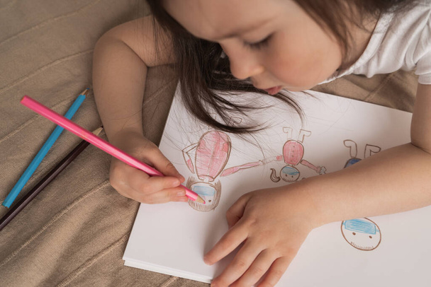 Cute girl rysuje rysunek ołówka. Dziecko uczy się rysować ludzi. Dziecko leży na łóżku i rysuje różowym ołówkiem.. - Zdjęcie, obraz