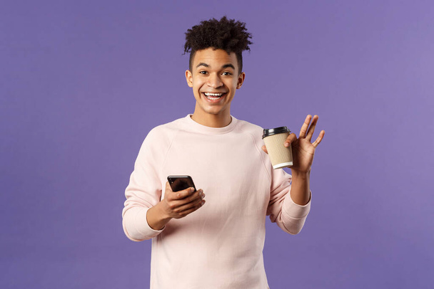 Porträt eines fröhlichen jungen männlichen Studenten mit Tasse Kaffee und Handy, winkende Hand zum Hallo sagen, freundlich lächelnd zur Begrüßung, Kommilitonen in der Nähe des Universitätscampus treffen, lila Hintergrund - Foto, Bild