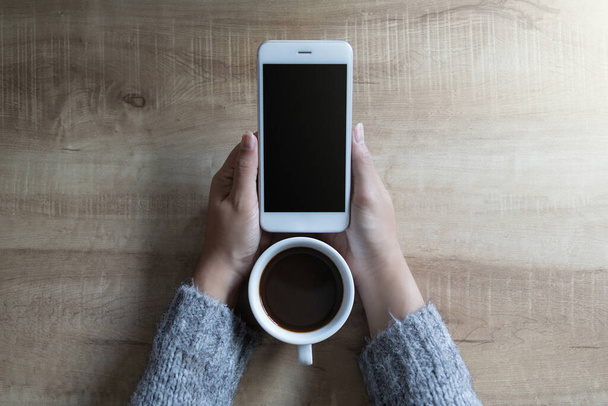 Attrappe der Hände einer Frau, die ein weißes Mobiltelefon mit leerem schwarzen Bildschirm und weißen heißen Kaffeetassen auf einem Holztisch im Café hält. - Foto, Bild