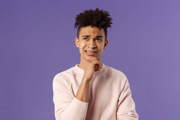 Close-up portrait of indecisive, puzzled young man facing difficult choice, look up thoughtful, touch chin and grimacing while thinking, making-up idea or decision, purple background - Photo, Image