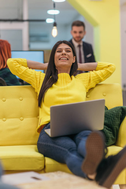 Bella donna d'affari (dipendente) seduta su un divano giallo mentre lavora sul suo computer portatile in startup. - Foto, immagini