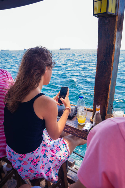 Brunette taking pictures with her smartphone of the beautiful sea while sitting in a bar on a boat - Photo, Image