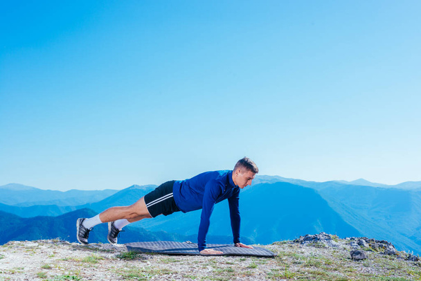 Strong handsome blonde athlete doing push ups on top of and mountain while enjoying the amazing view at the lake down in the valley. - Photo, Image