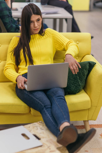 Όμορφη επιχειρηματίας (υπάλληλος) κάθεται σε ένα κίτρινο καναπέ, ενώ εργάζεται για το laptop της στην εταιρεία εκκίνησης. - Φωτογραφία, εικόνα