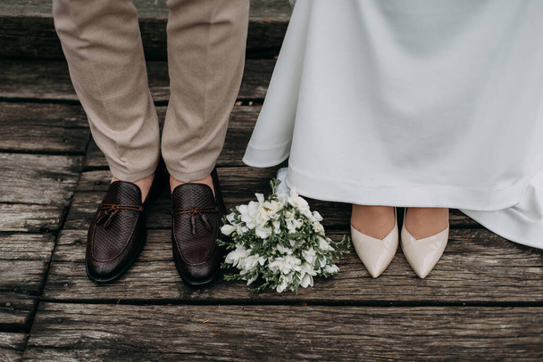 legs of newlyweds on wooden background. stylish shoes of bride and groom outdoors. bridal bouquet on brige close up. Stylish women's and men's shoes . wedding day. wedding details. wedding background - Photo, Image