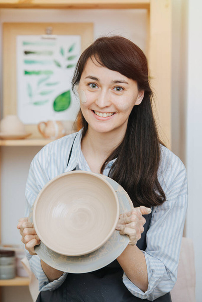 Master of ceramics in the studio. Portrait of a smiling woman with a clay bowl in her hands - Фото, изображение