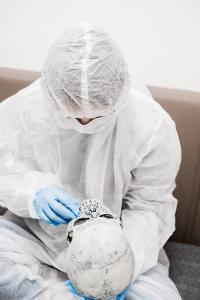 Hombre en traje blanco translúcido protector, guantes de goma azul, máscara médica se sienta en el sofá y sostiene el cráneo en sus manos delante de su cara. Permanecer en casa durante la pandemia del coronavirus
. - Foto, Imagen