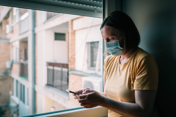 Woman in self-isolation during virus outbreak using mobile phone. Worried female person with protective surgical mask, holding smartphone, selective focus - Photo, image
