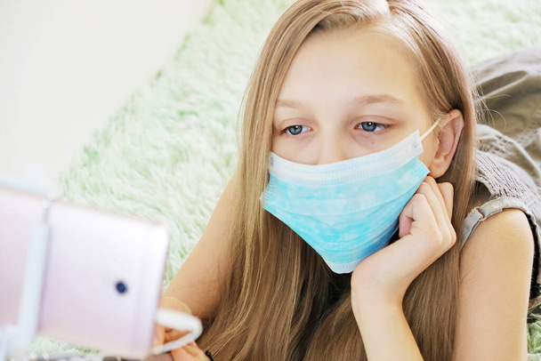 Girl puts on a preventive medical mask, has fun at home, taking pictures on the phone with help of facial gestures concept of home quarantine during global infectious coronavirus quarantine - Photo, image