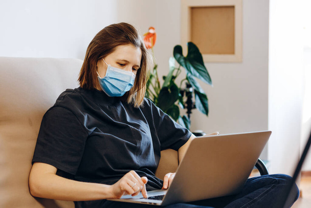 Evde dizüstü bilgisayarlı koruyucu maske takan bir kadın. Karantina, sosyal mesafe, evde kalmak, ofis kavramı. Coronavirüs salgını.. - Fotoğraf, Görsel
