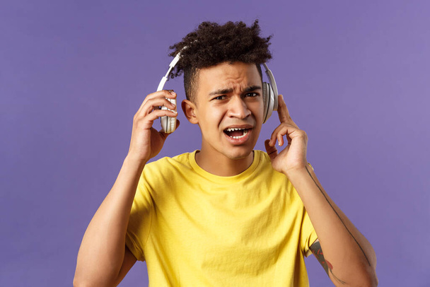 Cant hear you, repeat please. Portrait of young bothered guy interrupted of listening music, take-off headphones to answer person question, squinting look confused, purple background - Photo, Image