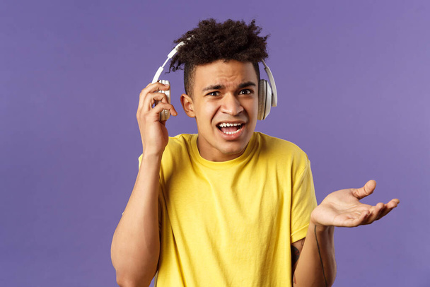 What do you want, I am in headphones. Portrait of confused annoyed young man shrugging, take-off earphone to hear what person asked while he was listening music, purple background - Photo, Image