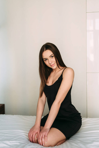 Portrait of a beautiful and sexy young adult attractive sensuality pretty brunette in elegant trendy black dress on the bed in the bedroom - Фото, изображение