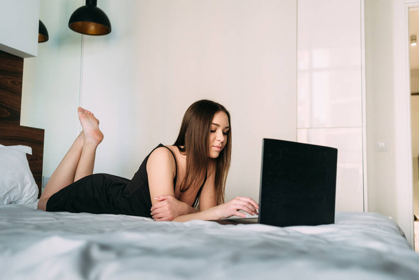 trabajo remoto en casa, una joven hermosa mujer se encuentra en casa en el sofá y trabaja en un ordenador portátil. Lugar para el texto
 - Foto, imagen