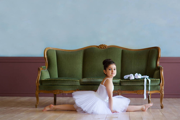 Mała dziewczynka balerina w białym tutu siedzi na podłodze w klasie baletu. - Zdjęcie, obraz