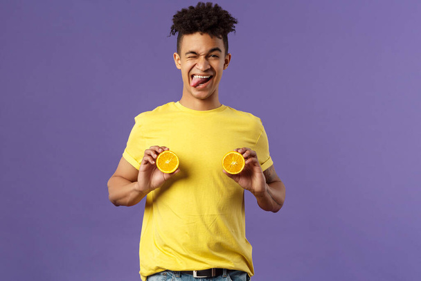 Holidays, vitamins and vacation concept. Portrait of sassy and cheeky funny young playful guy, fool around with fruit, holding pieces of oranges like women breast, show tongue smiling - Photo, Image
