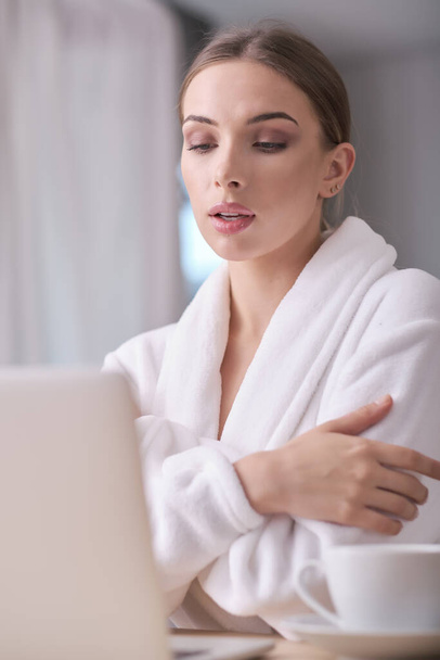 Unidentified young woman in a white bathrobe and towel sits on a cozy couch after bath surfing the Internet watches social networks using a laptop and high-speed Internet. Weekend at home concept - Foto, Imagem