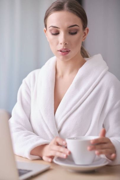 Unidentified young woman in a white bathrobe and towel sits on a cozy couch after bath surfing the Internet watches social networks using a laptop and high-speed Internet. Weekend at home concept - Foto, Imagem