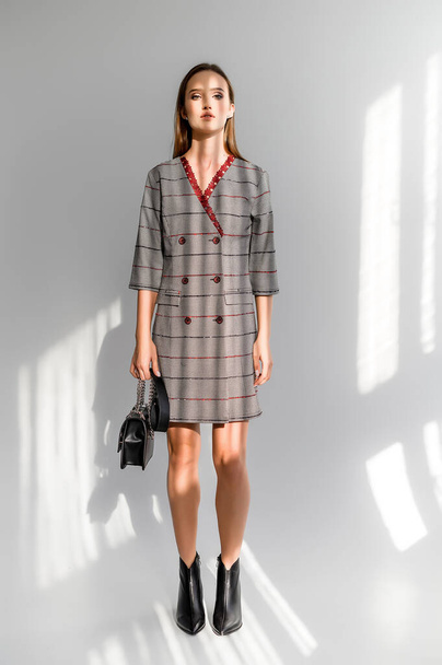 Attractive brown-haired woman in a gray checkered dress on a white background. Photosession for a clothing catalog - 写真・画像
