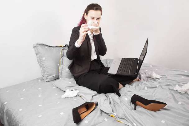 Concept - work from home, self-isolation, quarantine. Young girl in black suit sits in bed blow your nose and measuring temperature, holding laptop on her lap - Фото, изображение