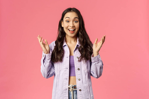 Portrait of surprised, enthusiastic korean girl in denim jacket, look wondered and happy, hear great news, applause, raise hands to clap and praise great work, smiling joyfully, pink background - Photo, Image