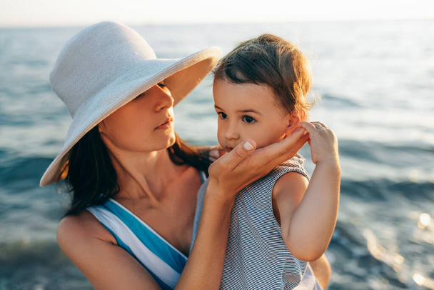 Portrait of candid beautiful mother care her cute daughter, wearing white hat and striped dress posing at the sea. Happy female with little girl walking at the ocean. Female with baby posing at ocean - Fotoğraf, Görsel