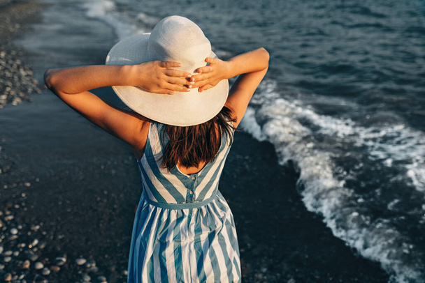 Closeup rear view image of gorgeous woman with white hat, walking along the beach at seaside on the sunset background. Female wearing striped dress posing outside during vacation. Travel concept.  - Фото, изображение