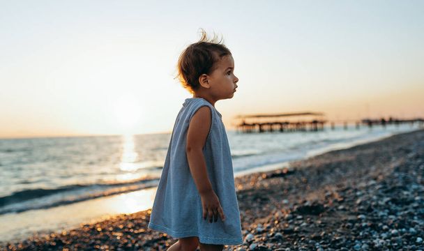 Horizontal image of the cute little girl wearing a dress walking at the sea shore on sunset background. Beautiful baby girl playing outside at the ocean beach on sunset. Childhood and travel concept - Foto, Imagen