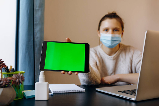 Jonge vrouw met een zwart tablet apparaat met groen scherm. Een vrouw die thuis werkt met een beschermend masker. Quarantaine, sociaal isolement, thuisblijven concept. - Foto, afbeelding